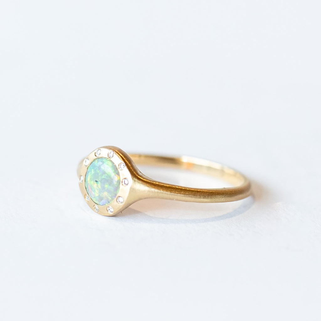 A matte-finished yellow gold dainty signet  ring featuring a round opal flush set in the face, surrounded by flush set white diamonds.