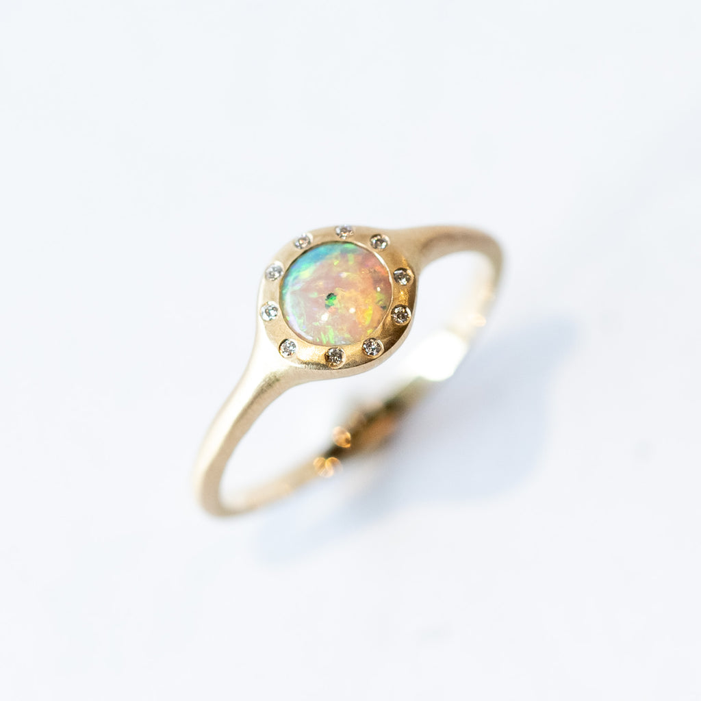 A matte-finished yellow gold dainty signet  ring featuring a round opal flush set in the face, surrounded by flush set white diamonds.
