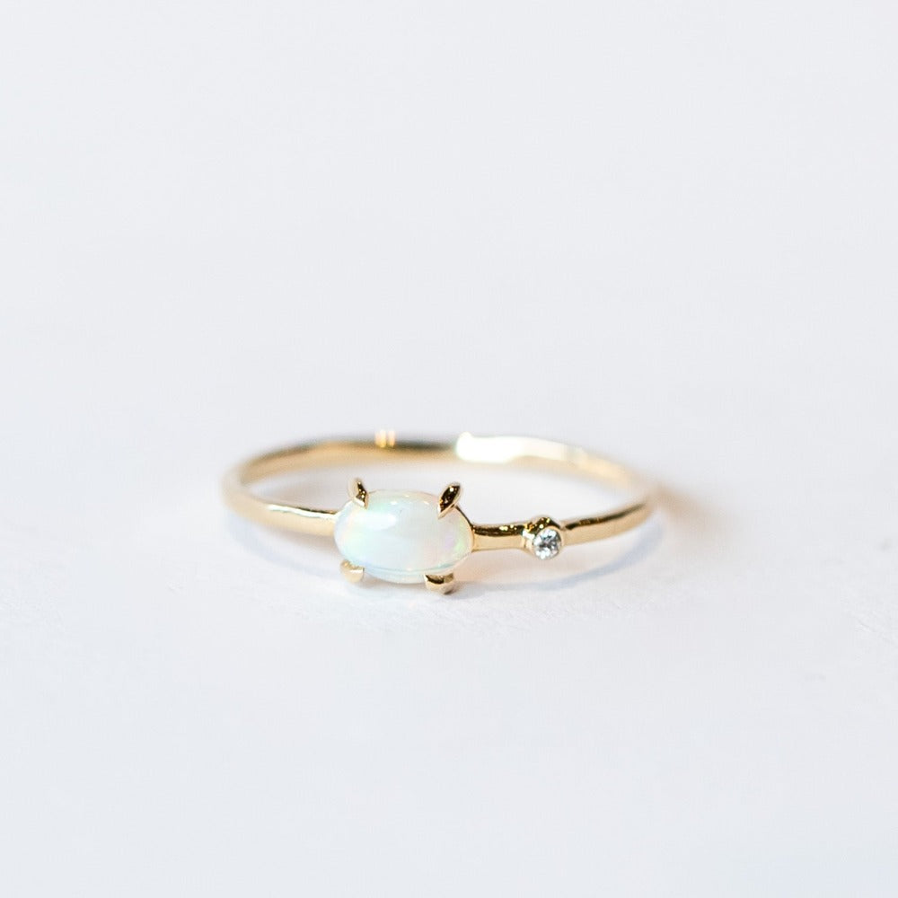 A thin yellow gold ring featuring a prong set oval cut white opal and a tiny bezel set round diamond to one side.