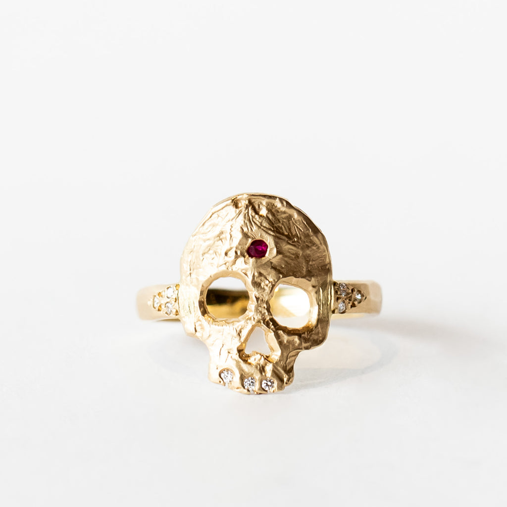 A hand carved gold ring with a skull shaped top, set with diamond teeth and a ruby on the forehead.