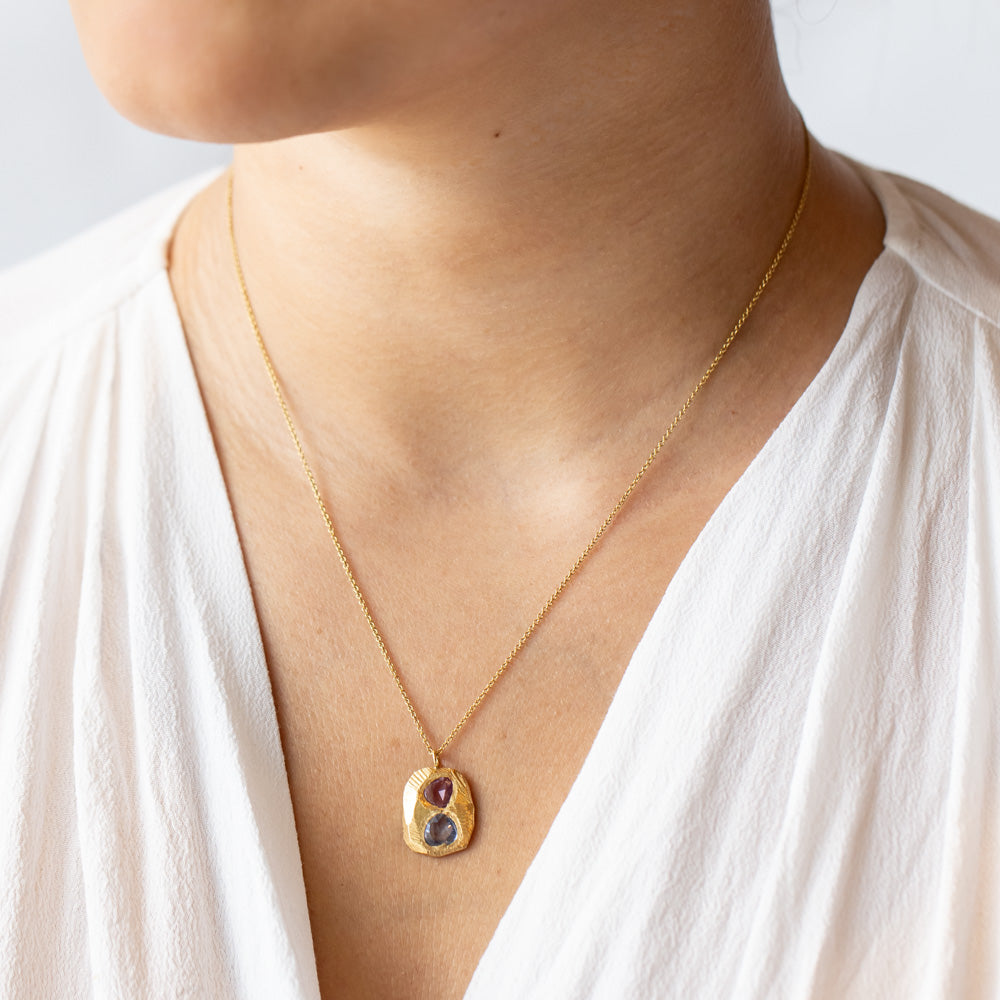 A carved yellow gold tablet is set with free form shaped sapphires, one pink, one purplish-blue on a cable chain necklace. On model.