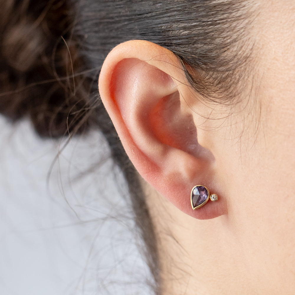 Pear shape purple sapphire stud earrings bezel set in yellow gold and accented by tiny round diamonds. On model.