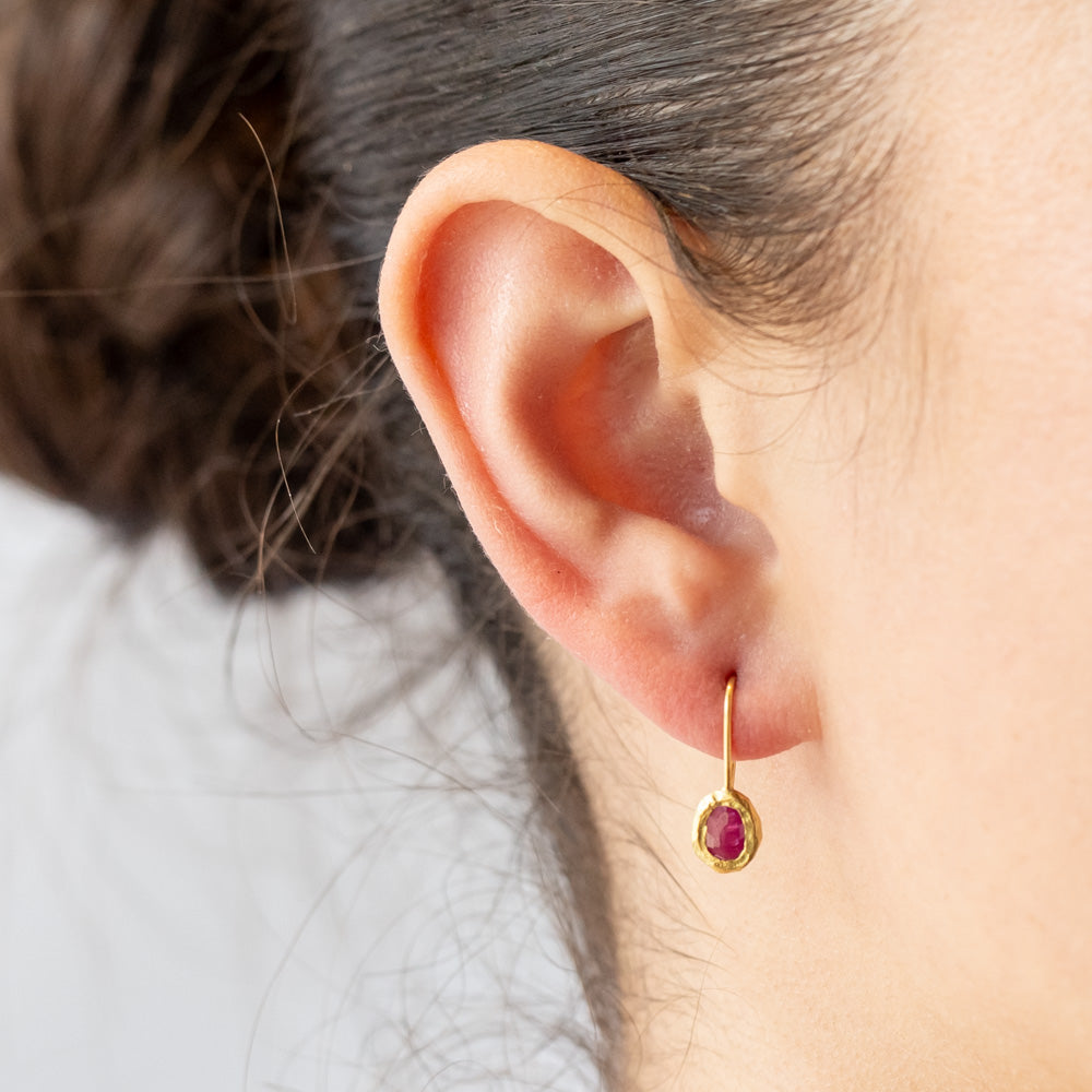 Dainty yellow gold drop earrings with oval rubies set in textured bezels. On model.