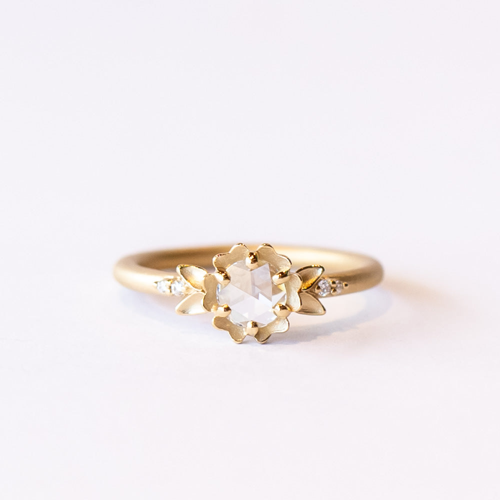 A yellow gold engagement ring featuring a round, rose cut white diamond set at the center of a carved buttercup flower. Each side is accented with leaves and tiny white diamonds.