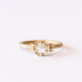 A yellow gold engagement ring featuring a round, rose cut white diamond set at the center of a carved buttercup flower. Each side is accented with leaves and tiny white diamonds.