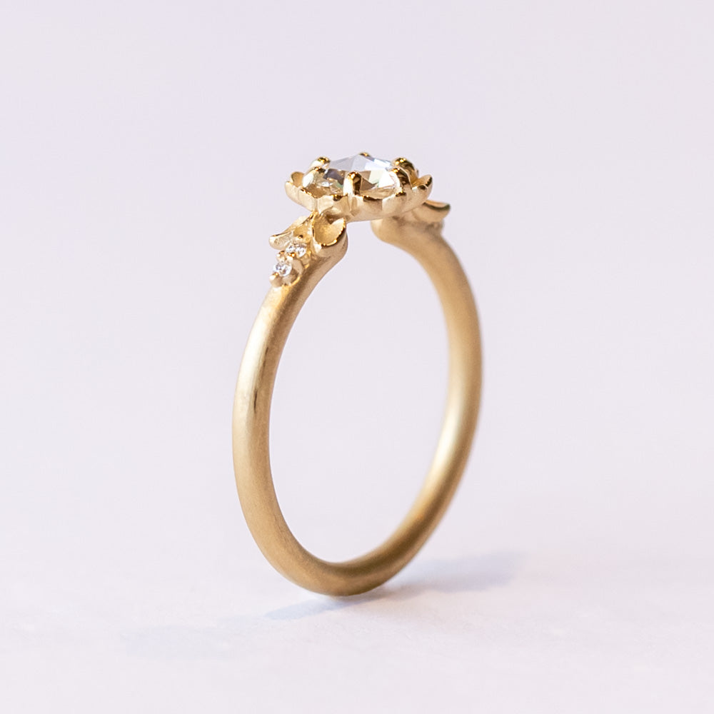 A yellow gold engagement ring featuring a round, rose cut white diamond set at the center of a carved buttercup flower. Each side is accented with leaves and tiny white diamonds.