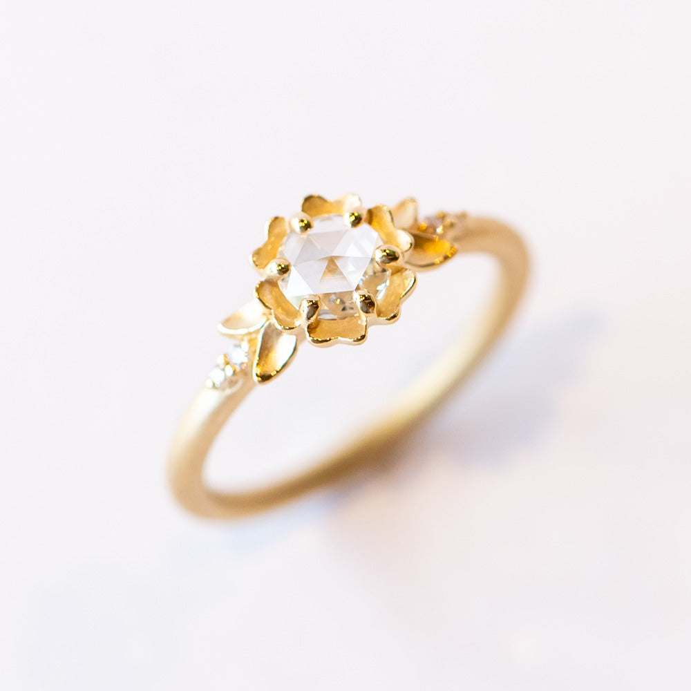 A yellow gold engagement ring featuring a round, rose cut white diamond set at the center of a carved buttercup flower. Each side is accented with leaves and tiny white diamonds.