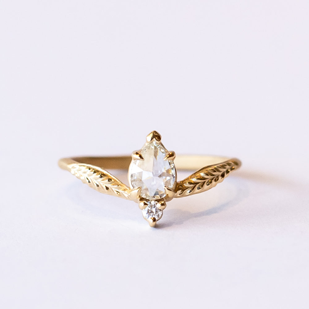 A unique yellow gold engagement ring featuring a peaked design set with an asymmetrical diamond paring of a pear shape rose cut with a tiny round diamond. Leaf accents adorn each shoulder.