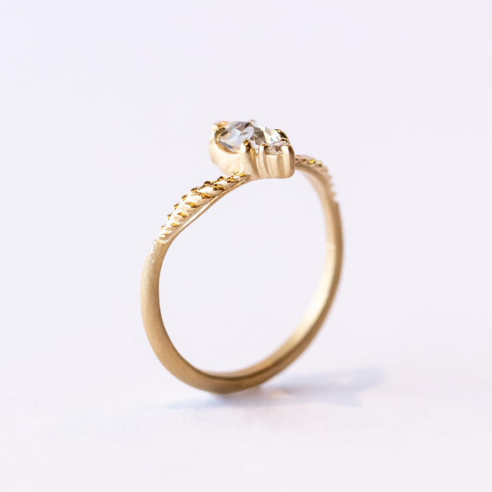 A unique yellow gold engagement ring featuring a peaked design set with an asymmetrical diamond paring of a pear shape rose cut with a tiny round diamond. Leaf accents adorn each shoulder.