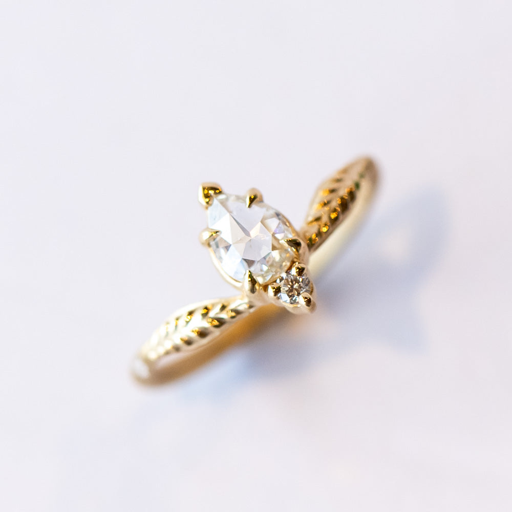 A unique yellow gold engagement ring featuring a peaked design set with an asymmetrical diamond paring of a pear shape rose cut with a tiny round diamond. Leaf accents adorn each shoulder.