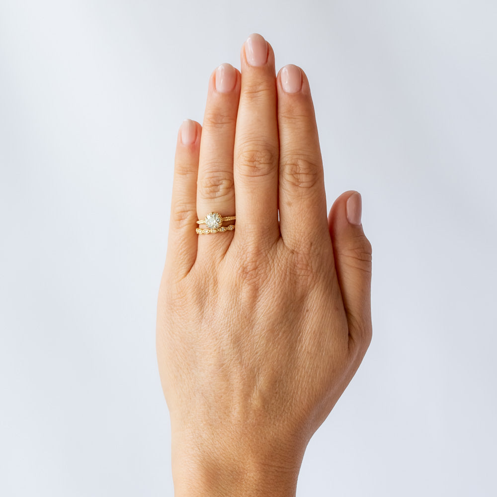 A yellow gold solitaire diamond engagement ring featuring a prong set old european cut diamond and a slender matte finish, leaf accented band. On hand with a wedding band.