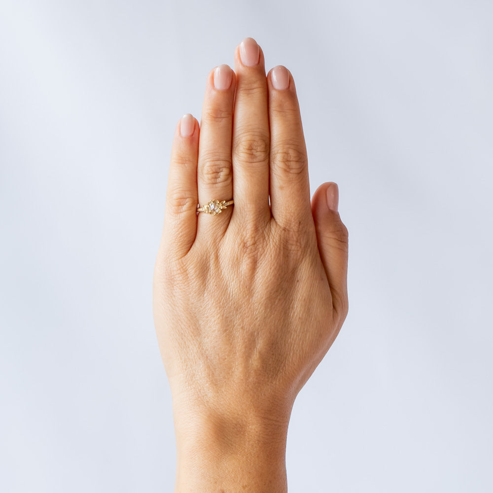 A yellow gold engagement ring featuring a round, rose cut white diamond set at the center of a carved buttercup flower. Each side is accented with leaves and tiny white diamonds. On hand.