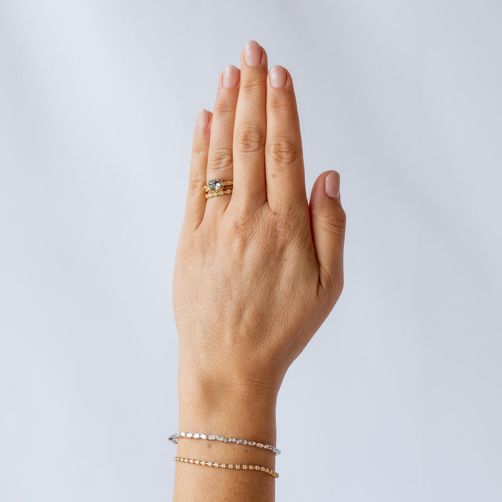 A cushion cut salt and pepper diamond is prong set into a yellow gold engagement ring. The sides of the setting feature an asymmetrical floral motif set with two tiny accent diamonds. On hand.