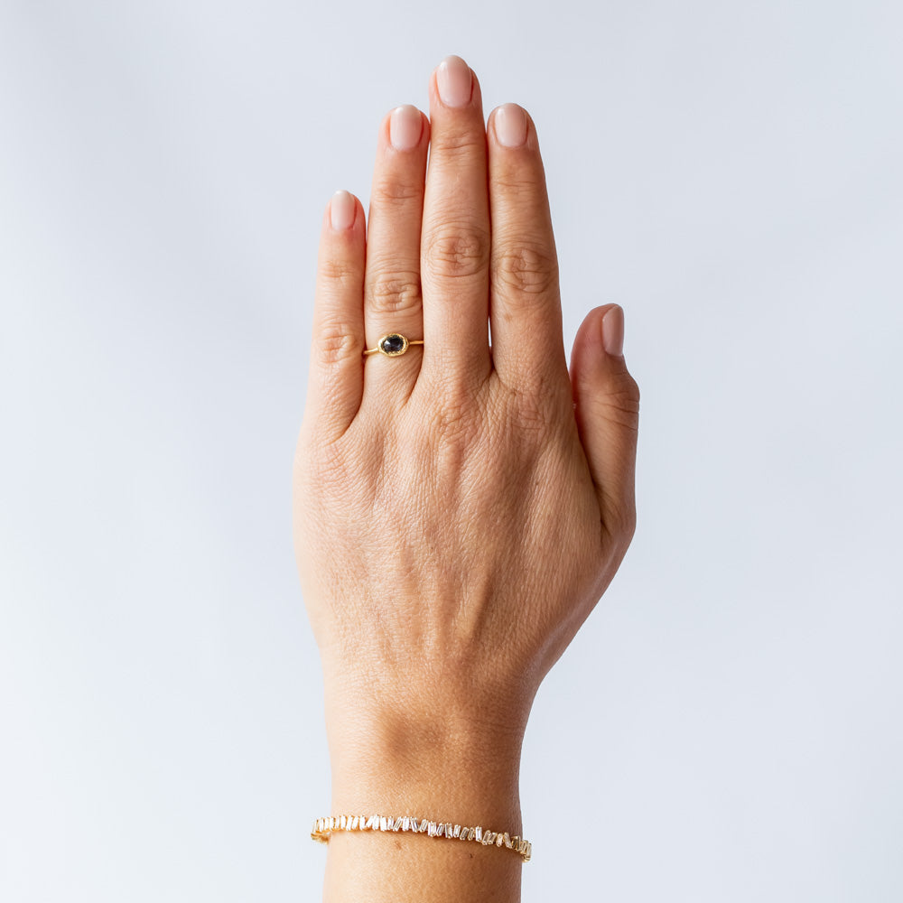 A slender, textured yellow gold ring with an east-west, bezel set teal blue sapphire. On hand.