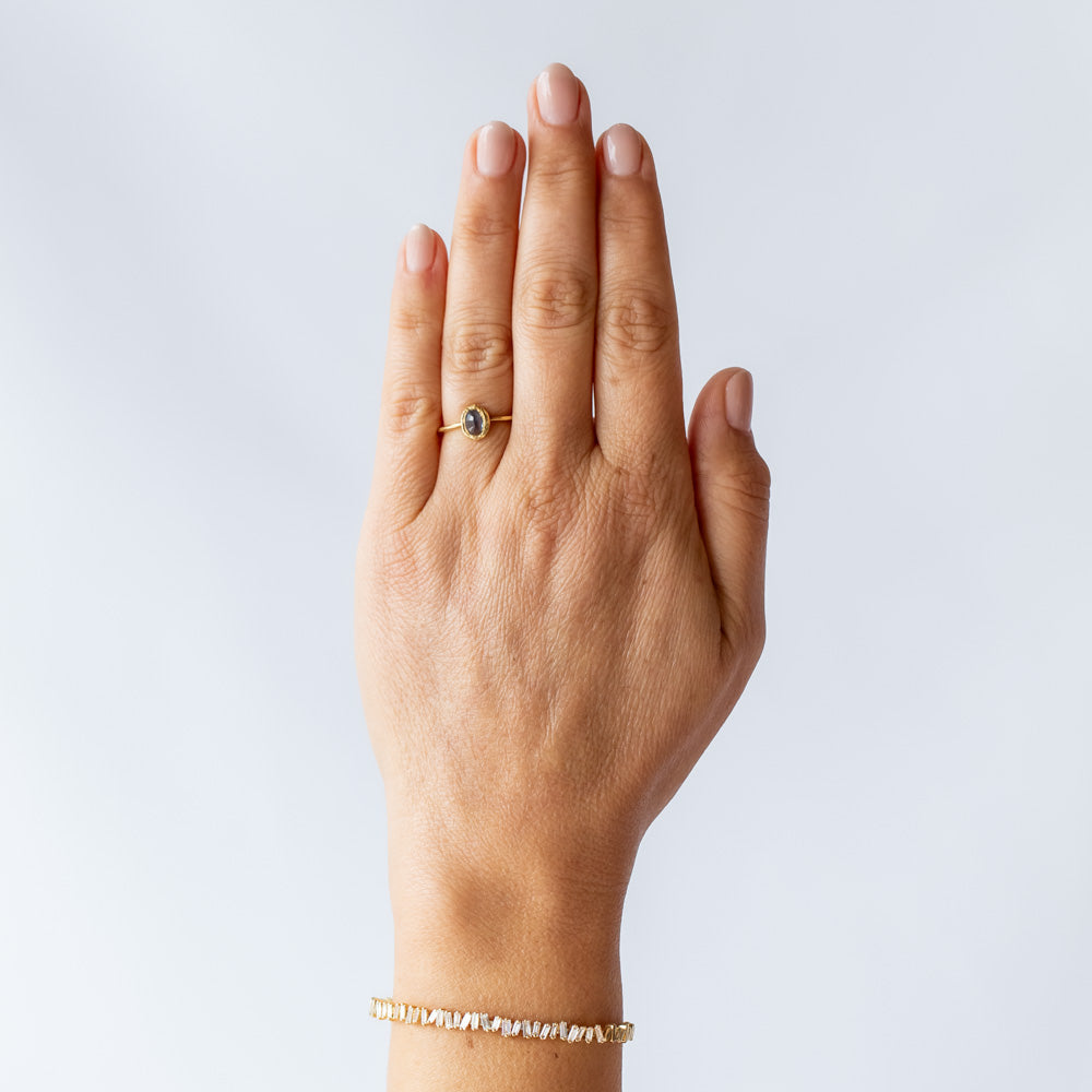 A petite yellow gold ring featuring hand carved texture and a bezel set oval rose cut blue sapphire. On hand.