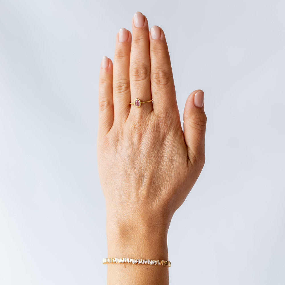 A oval rose cut pink sapphire is set into a textured bezel on a slender yellow gold ring. On model.