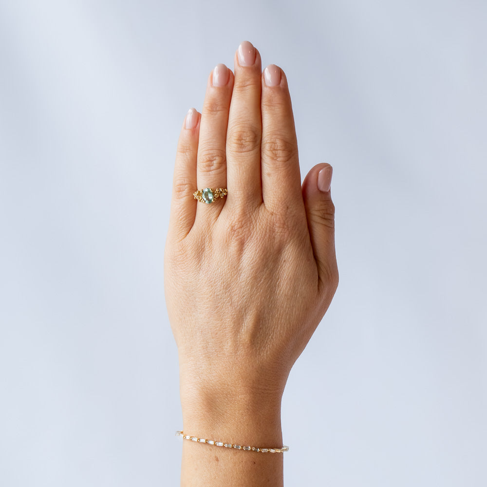 A pale blue-green oval tourmaline set in golden granules with diamond accents on a yellow gold ring. On hand.