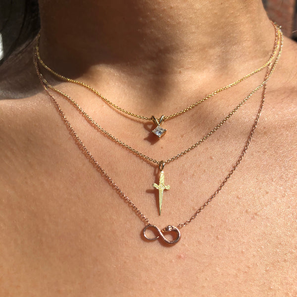 A tiny gold engraved dagger pendant necklace layered with other necklaces.