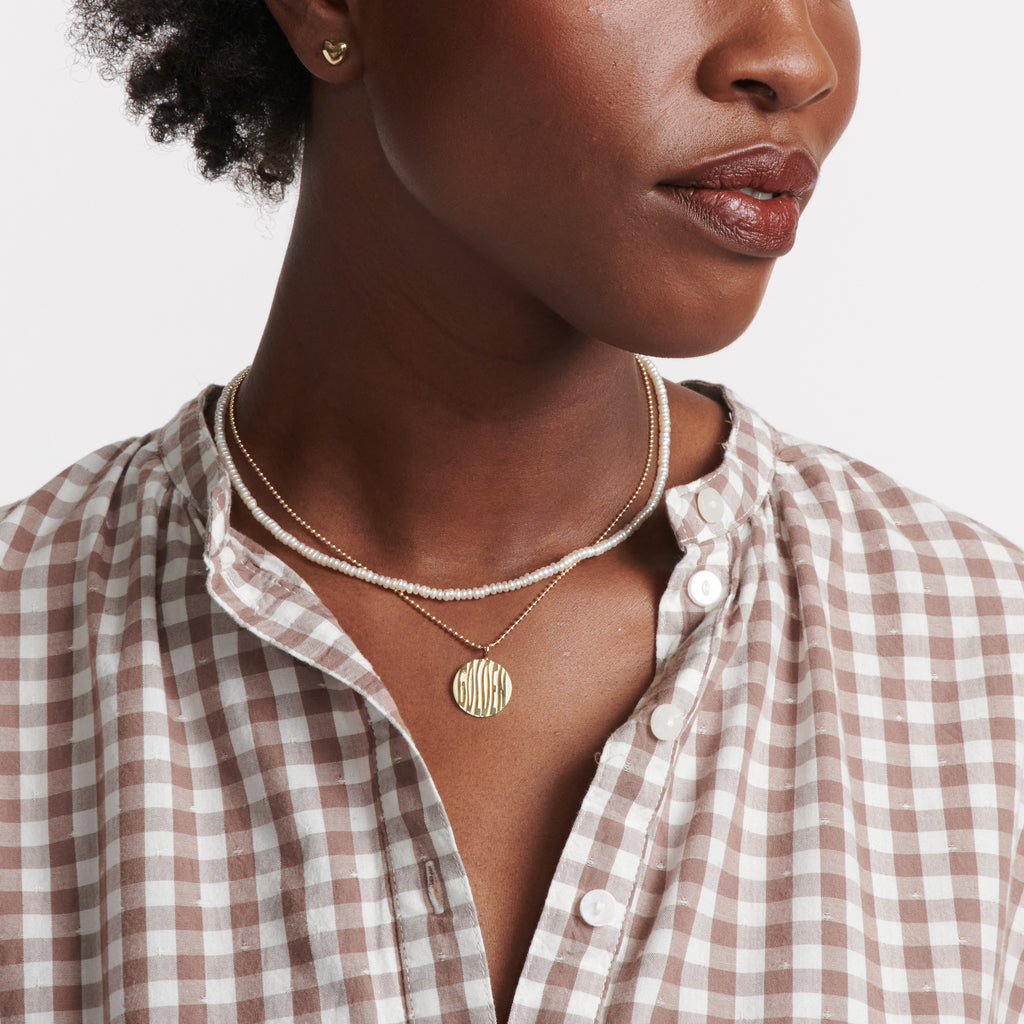 A round gold pendant featuring wavy letters spelling "golden" and suspended from a gold ball chain necklace. on model.