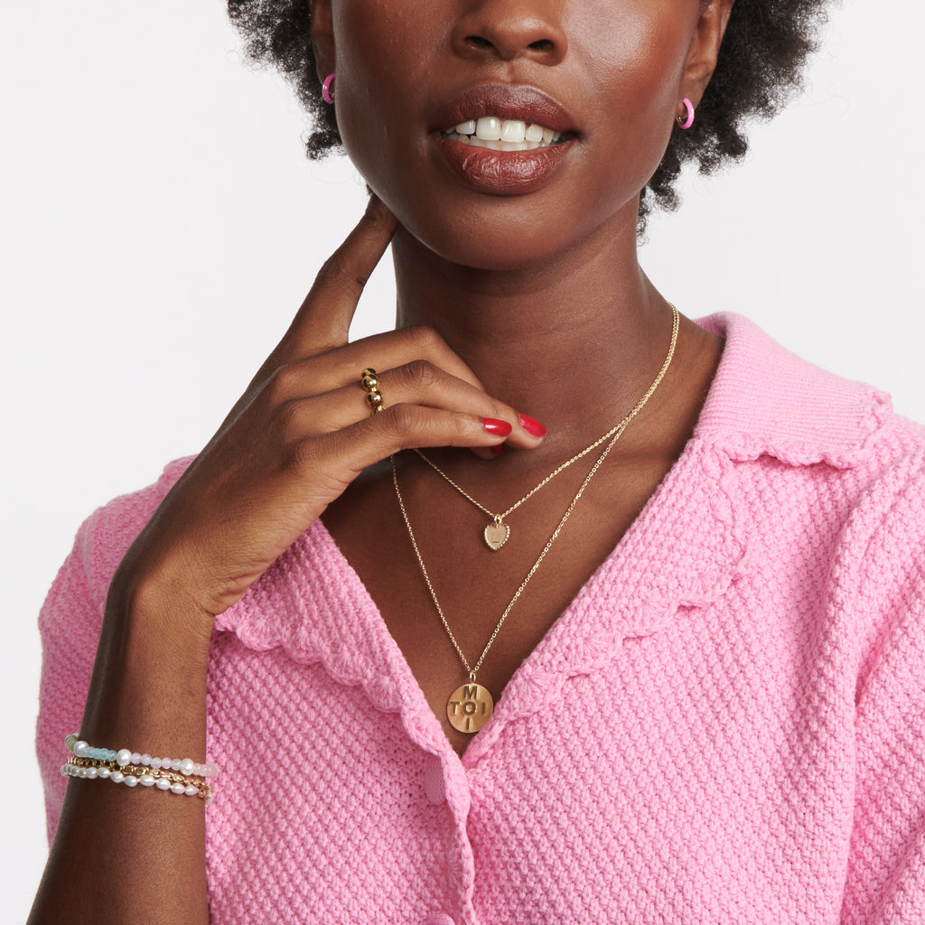 A round gold pendant with a carved starburst texture and the words "toi" and "moi" crossed in block letters through the middle. Hands from a simple gold chain necklace. On model.