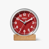 Shinola red desk clock with silver case, sitting on a tan wooden block, front view
