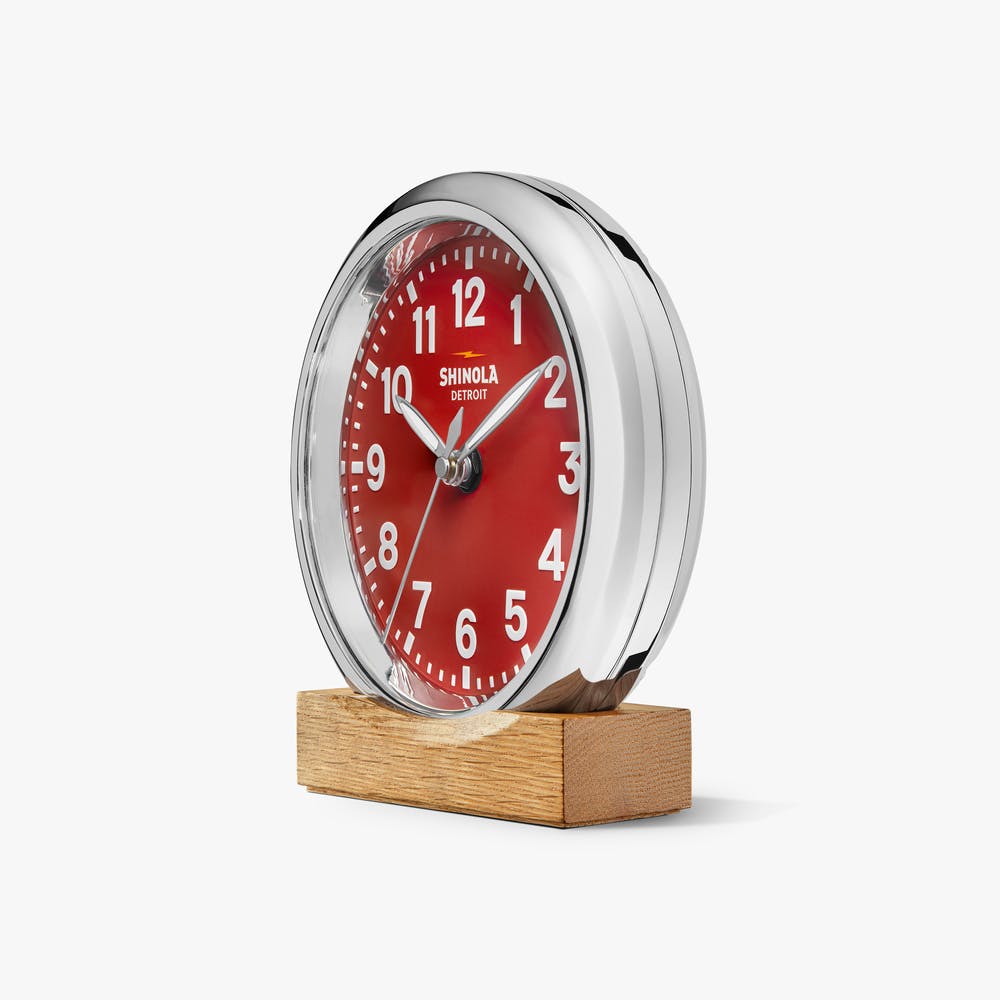 Shinola red desk clock with silver case, sitting on a tan wooden block, angled front view