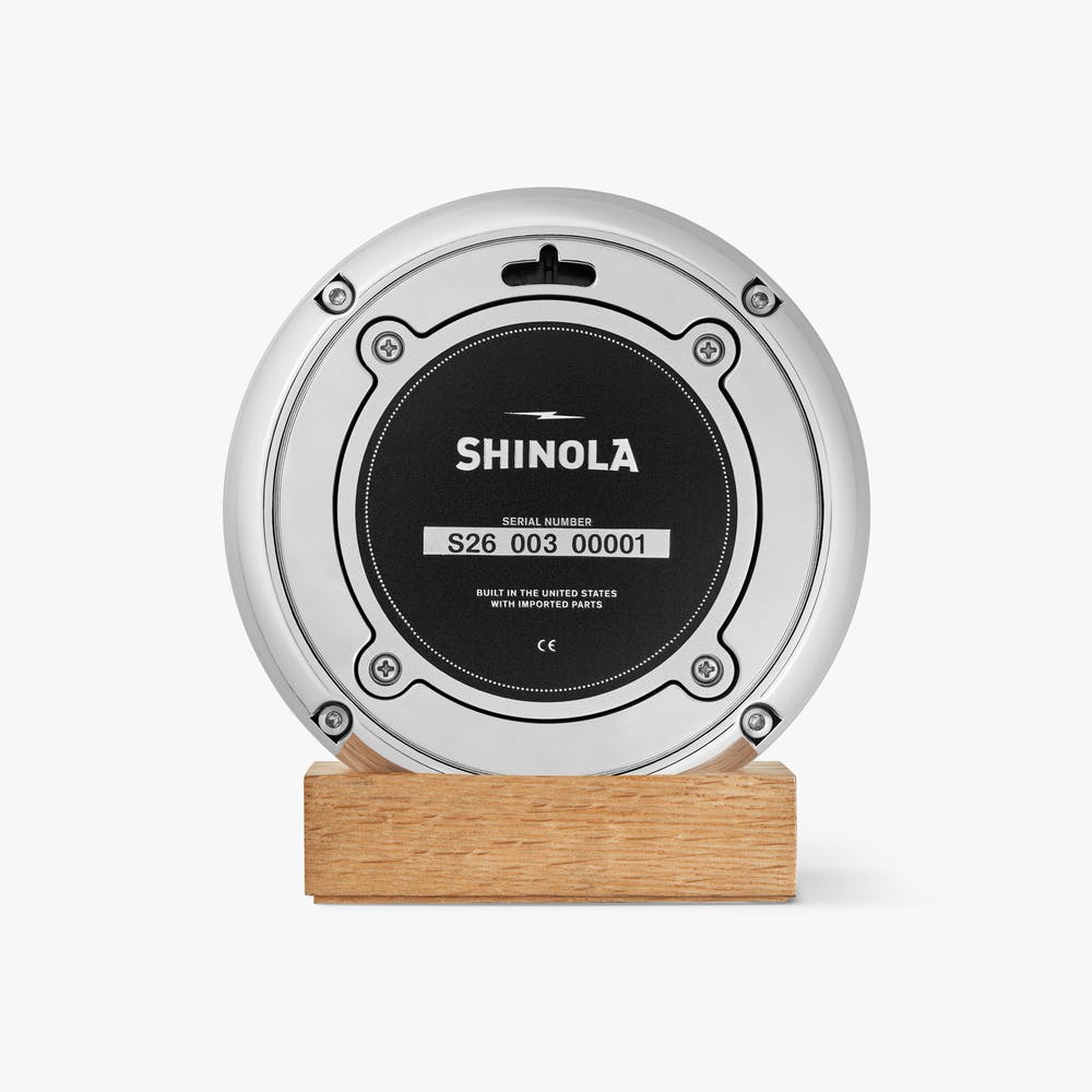 Shinola red desk clock with silver case, sitting on a tan wooden block, back view